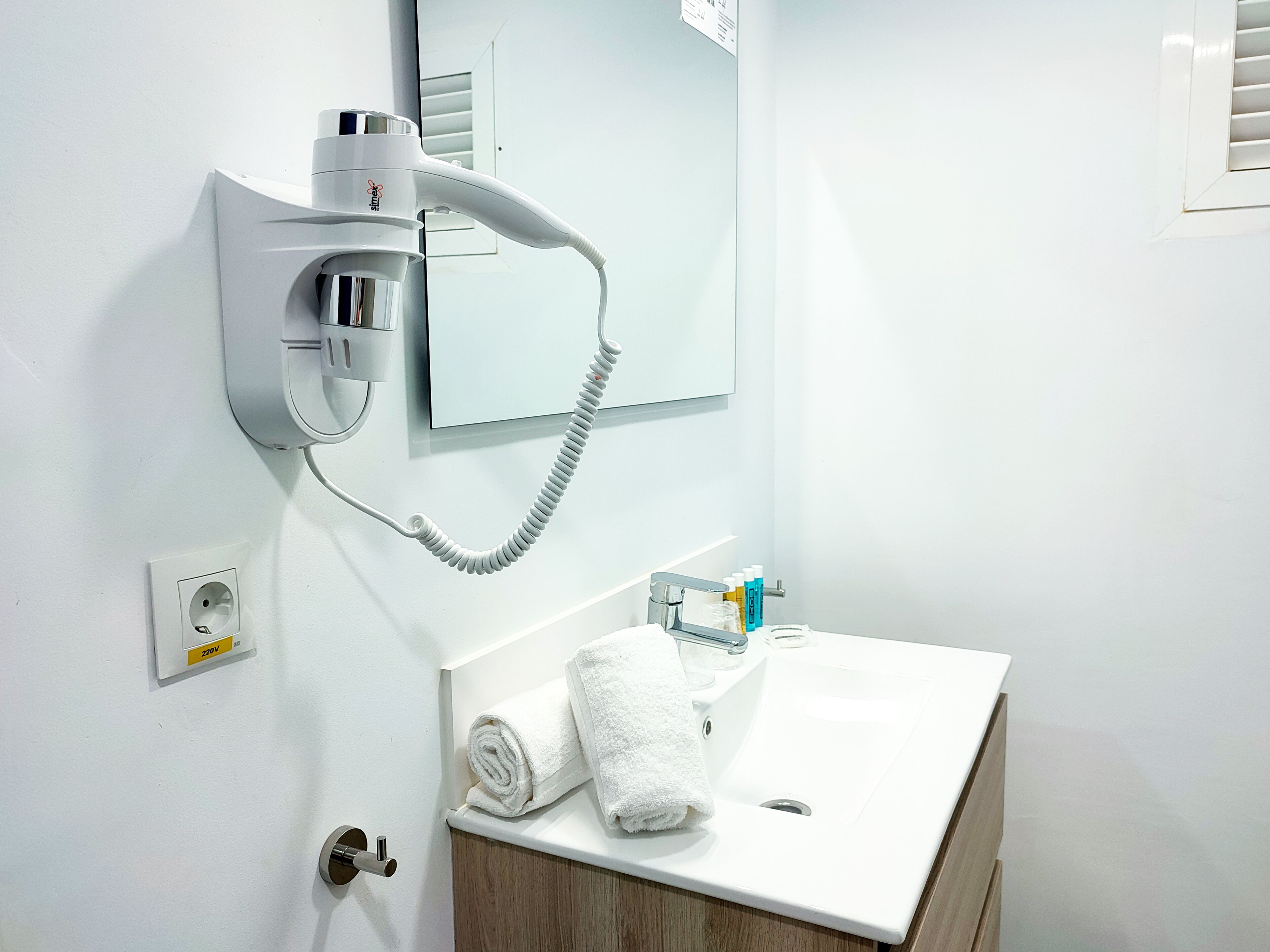 A modern bathroom with a hairdryer mounted on the wall, a sink with a mirror above it, and a towel rolled up on the counter.