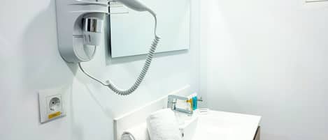 A modern bathroom with a hairdryer mounted on the wall, a sink with a mirror above it, and a towel rolled up on the counter.