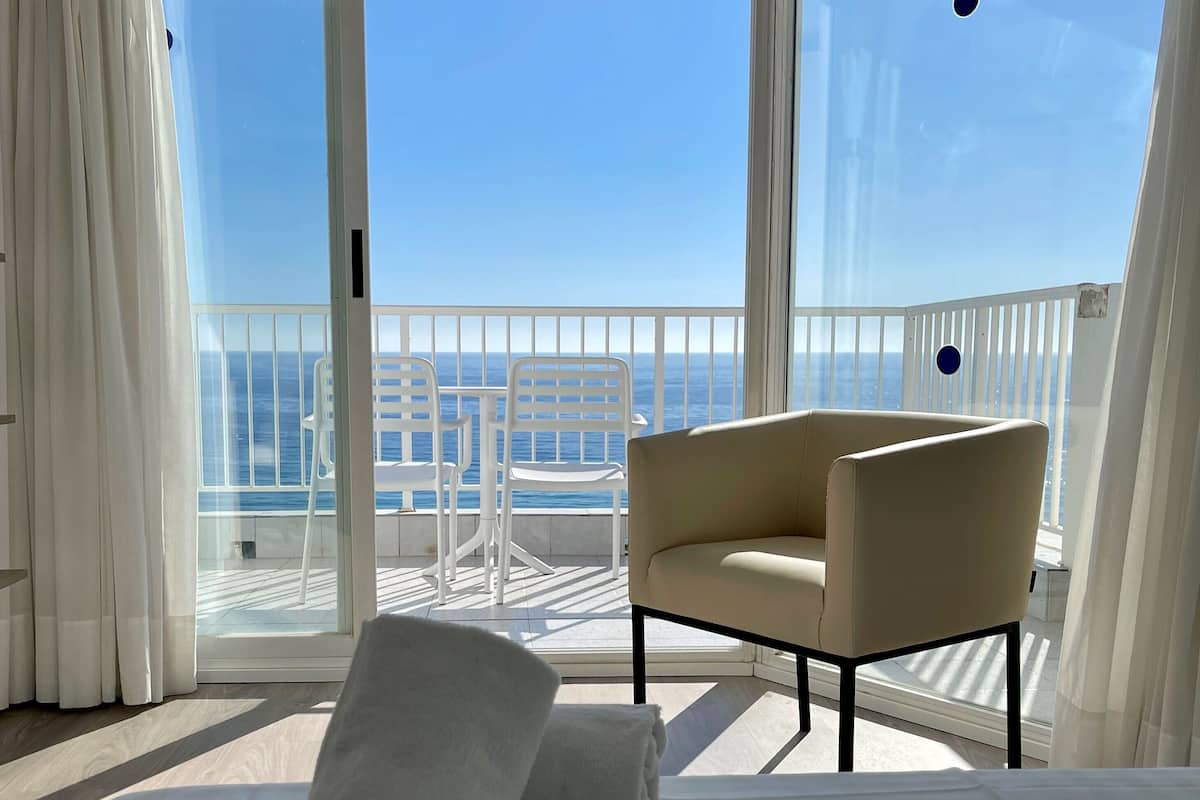 Una habitación con cama, una silla y vista al mar a través de amplios ventanales.