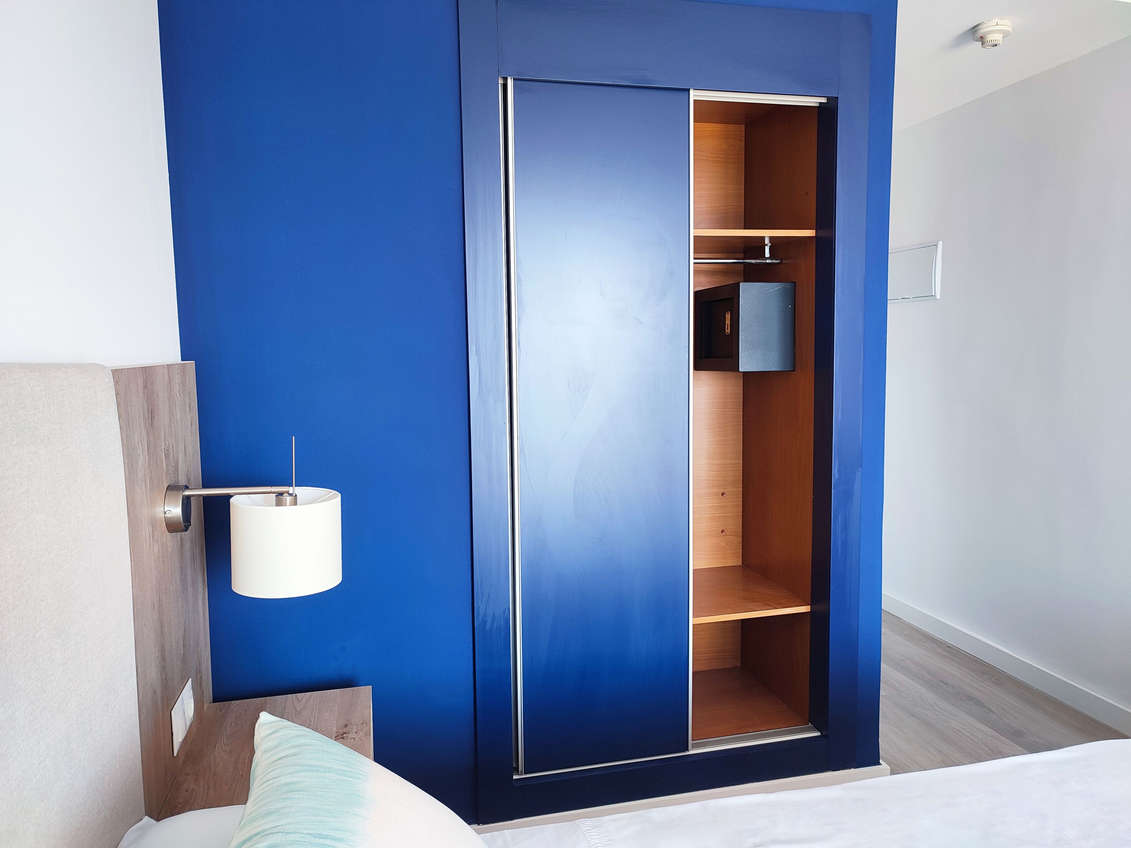 A bedroom with a blue wall, a wooden wardrobe, a bedside table with a lamp, and a bed with a white headboard.
