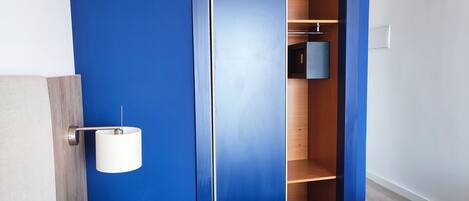 A bedroom with a blue wall, a wooden wardrobe, a bedside table with a lamp, and a bed with a white headboard.