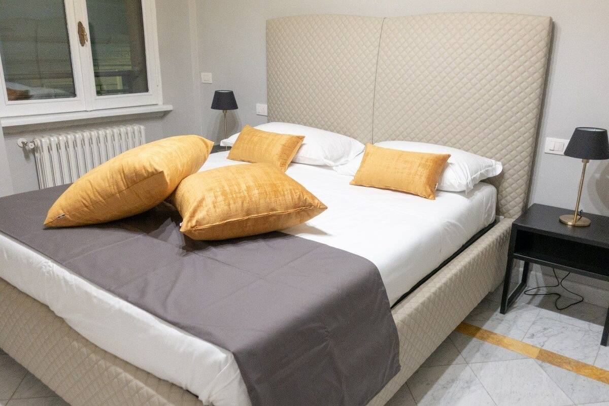A neatly made bed with a grey bedspread and multiple yellow pillows, a bedside table with a lamp, and a window with blinds.