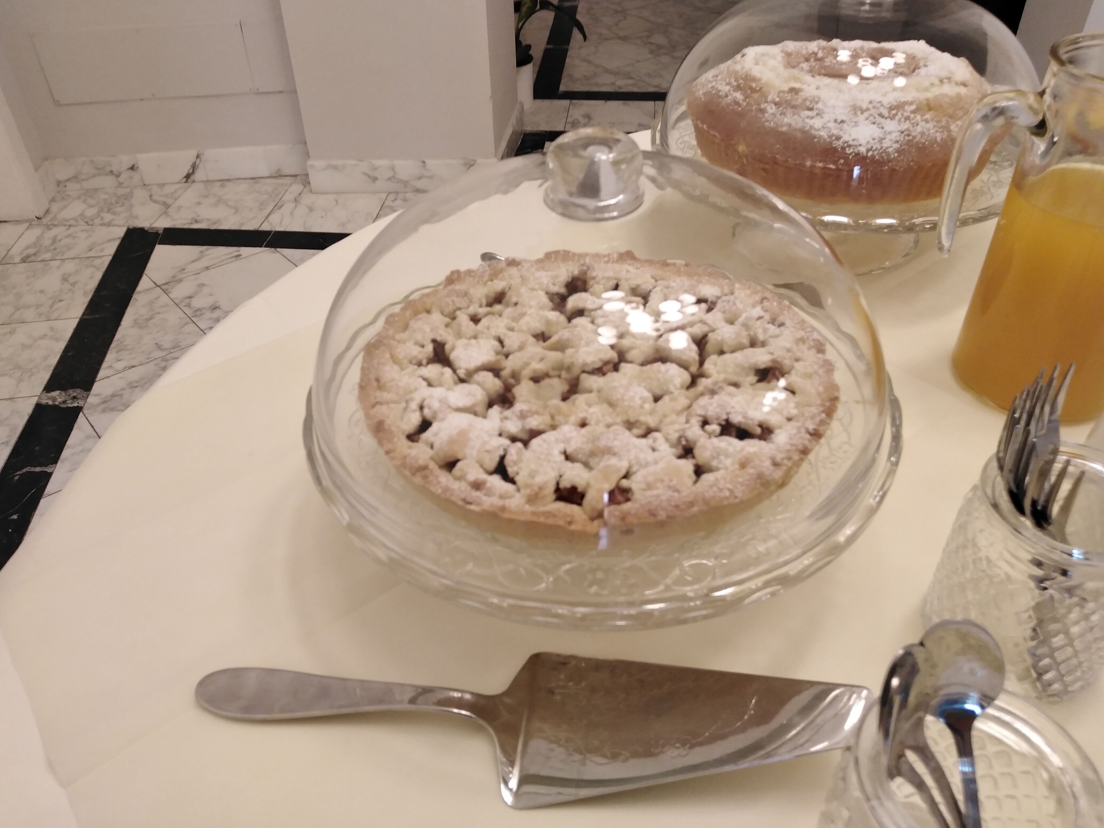 A dessert table with a covered pie, a jug of orange juice, and a pie server.