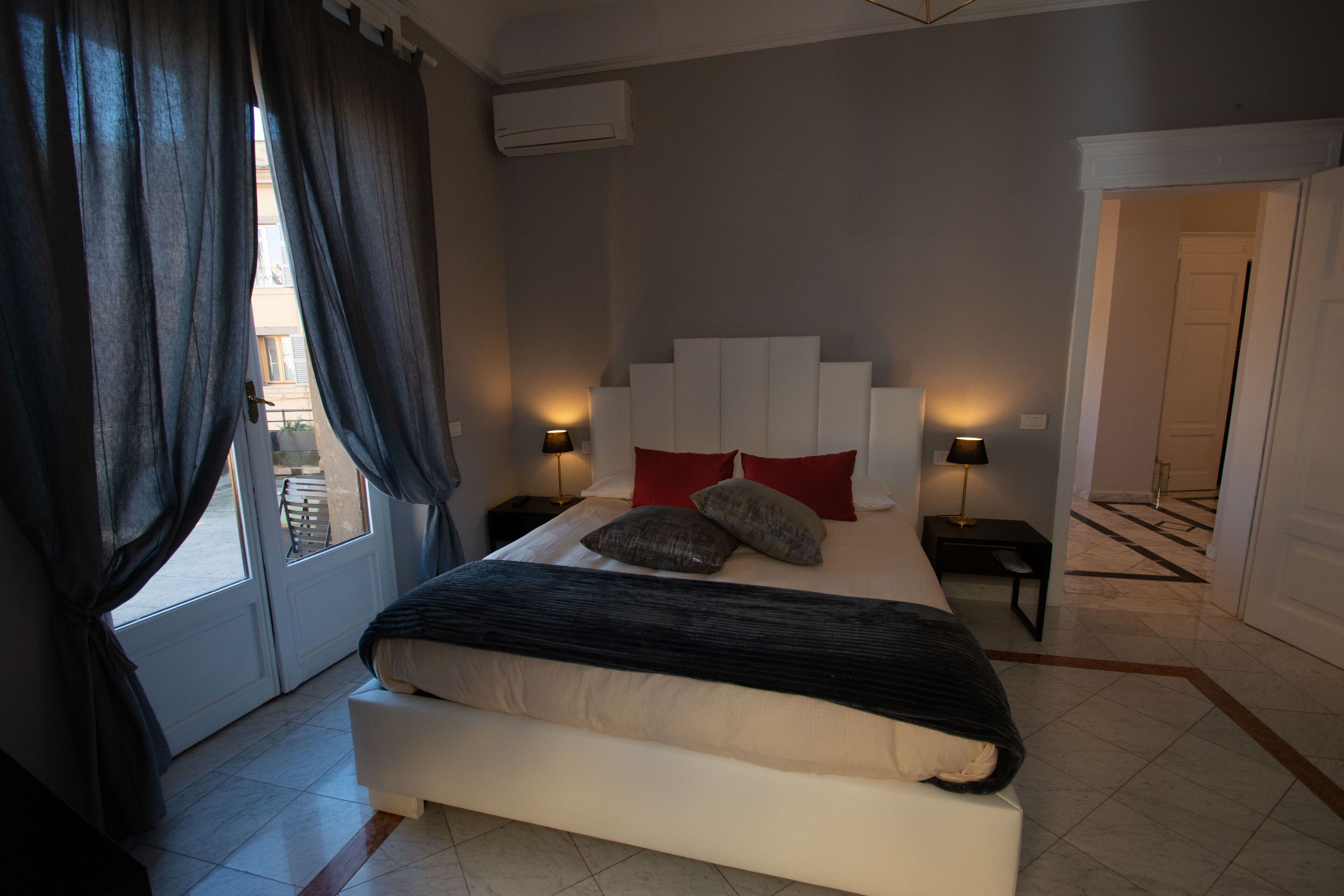 A bedroom with a large bed, two bedside tables with lamps, a window with curtains, and a door leading to another room.