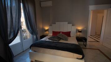 A bedroom with a large bed, two bedside tables with lamps, a window with curtains, and a door leading to another room.