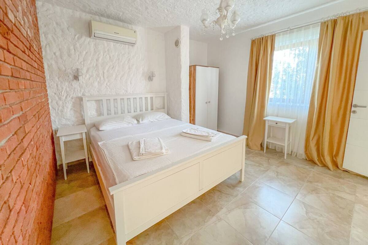 A bedroom with a white bed, a small bedside table, a mirror, and a window with curtains.
