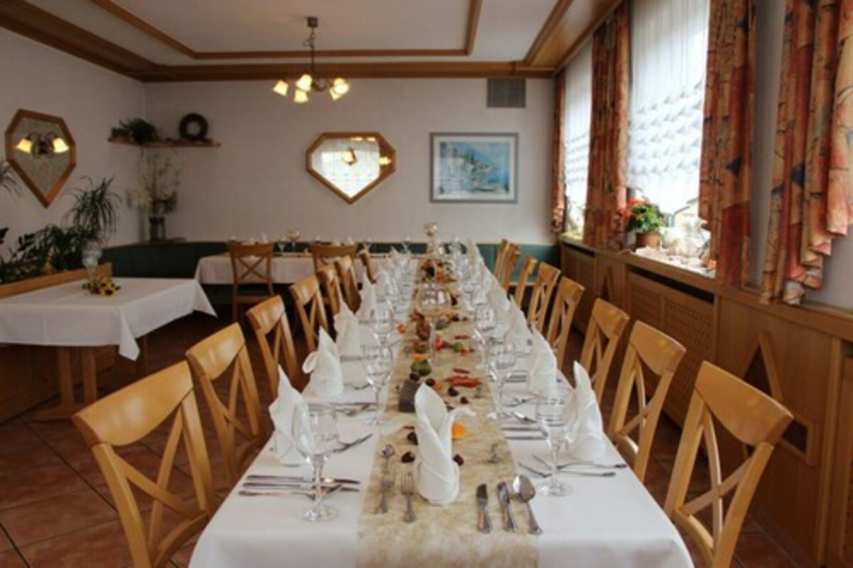 Une longue table à manger, recouverte de nappes blanches et entourée de chaises en bois, se trouve dans une pièce dont les murs sont ornés d’éléments décoratifs et qui possède une fenêtre agrémentée de rideaux.