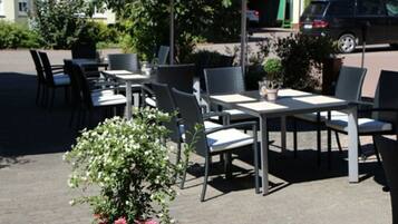 Een terras met tafels en stoelen, een plant in een pot en een geparkeerde auto op de achtergrond.