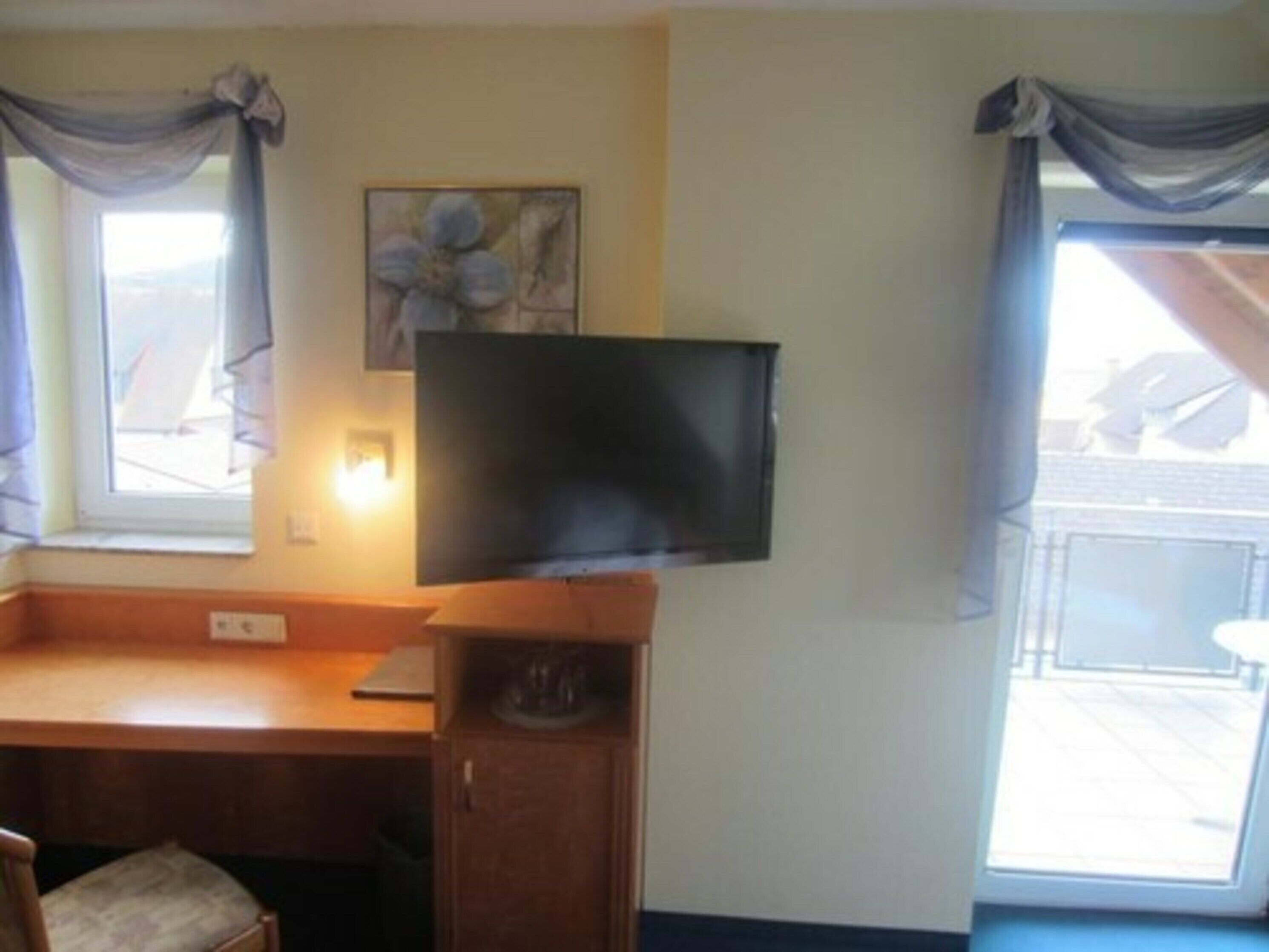 A room with a wooden desk, a television mounted on the wall, a window with curtains, and a painting on the wall.
