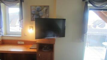 A room with a wooden desk, a television mounted on the wall, a window with curtains, and a painting on the wall.