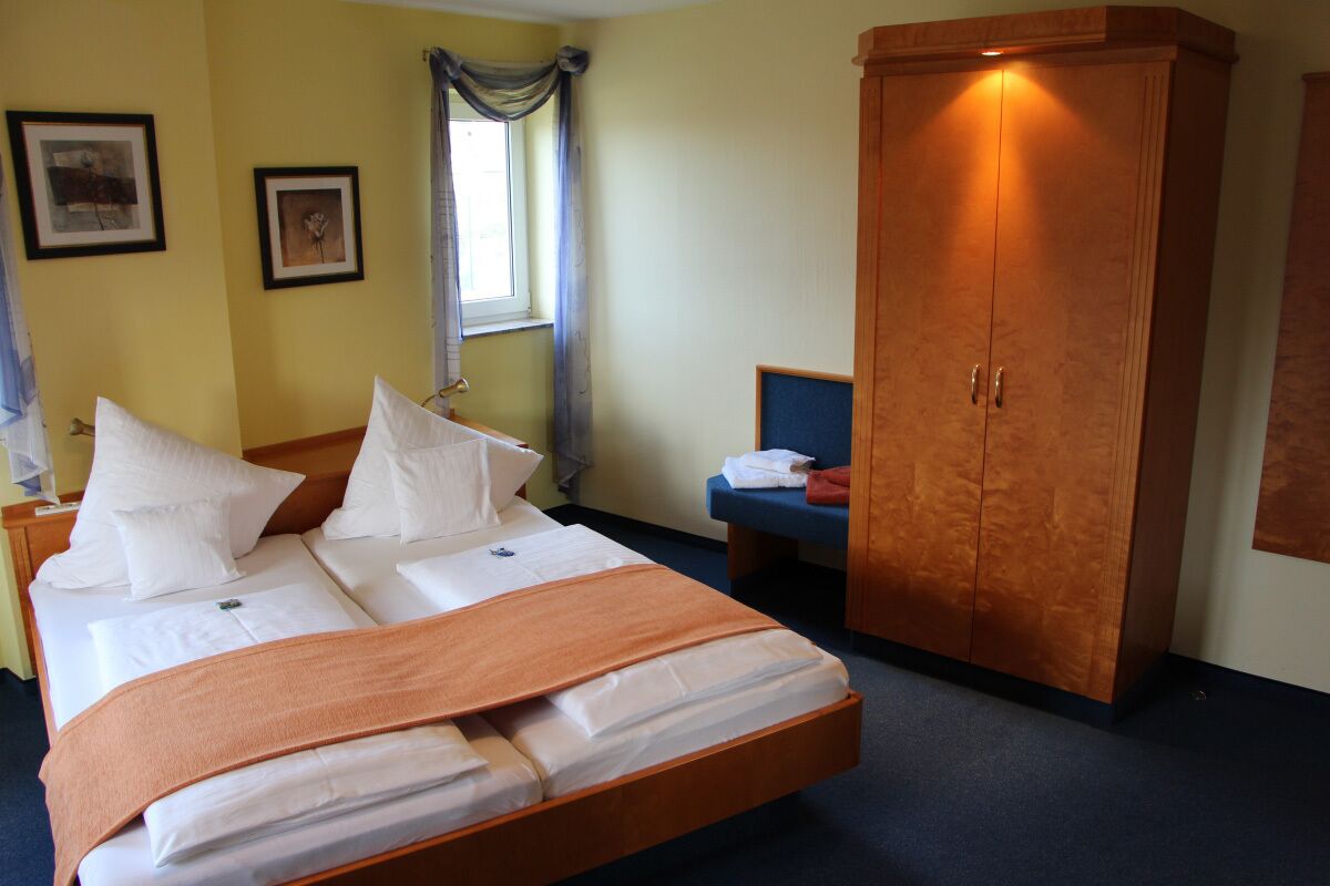A hotel room with a bed, a wardrobe, a window with curtains, and two framed pictures on the wall.