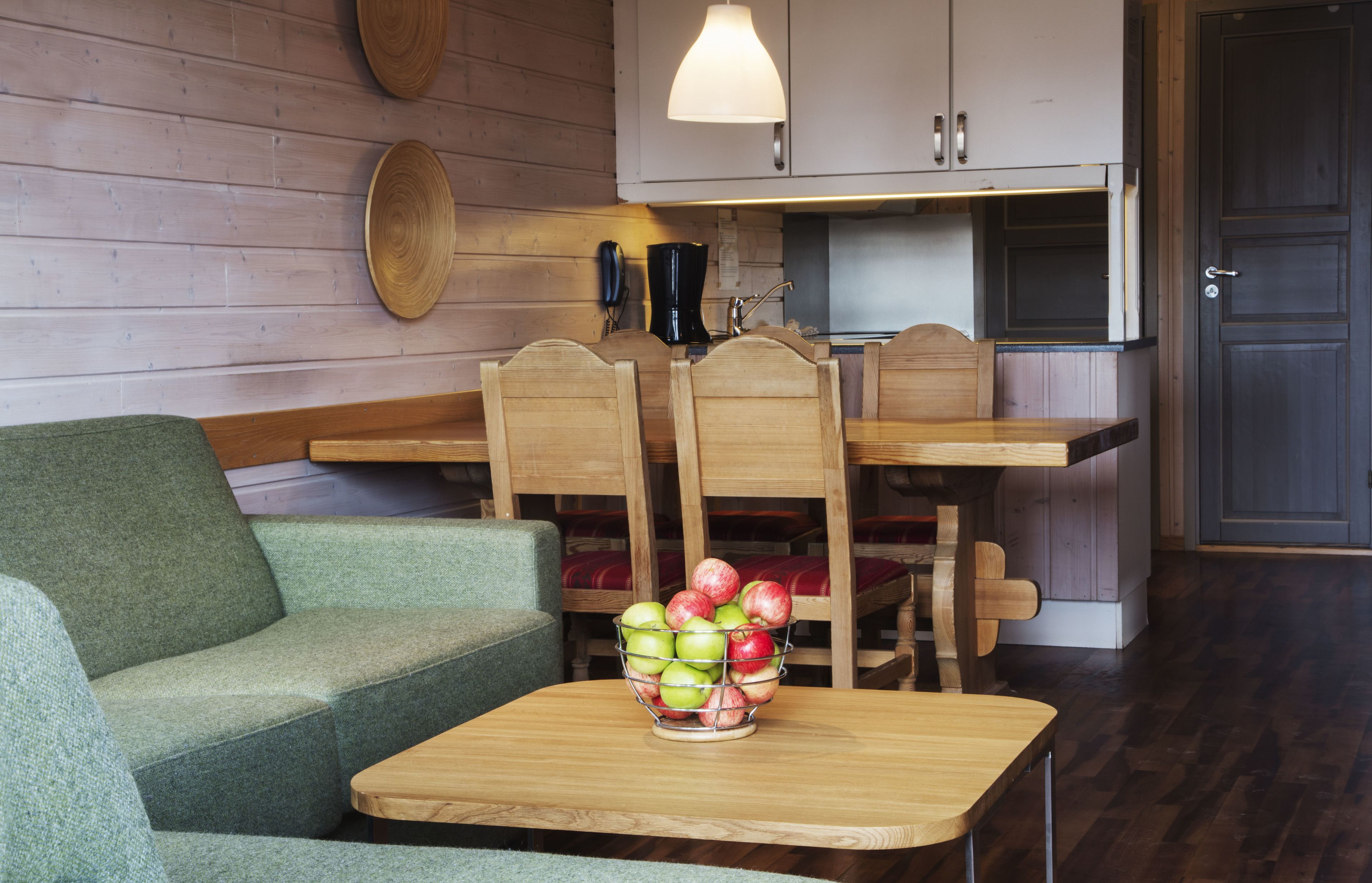 A cozy room with a green sofa, wooden dining table, and chairs. A fruit bowl is on the table.