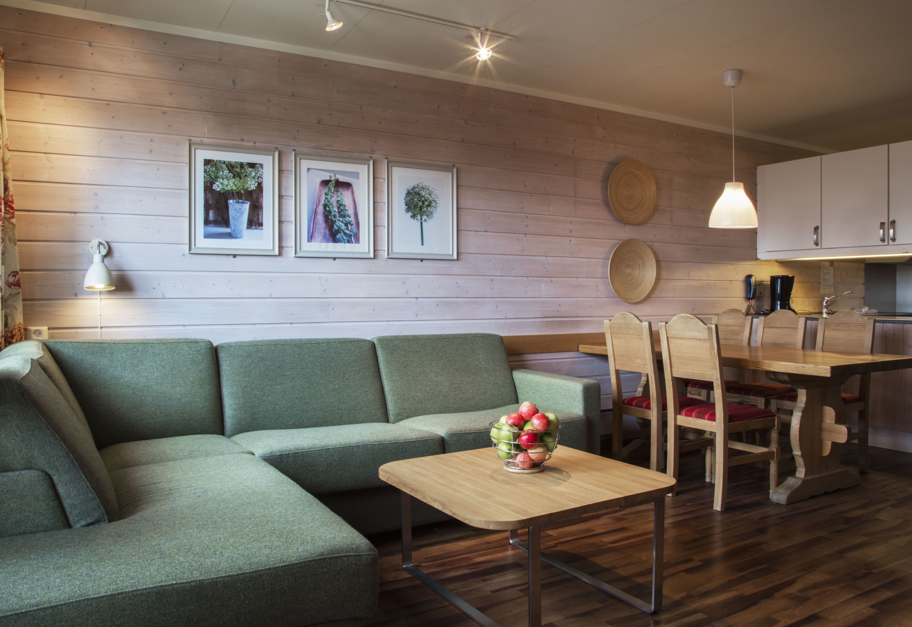 A modern living room with a green sofa, a wooden dining table with chairs, and a small wooden coffee table with a fruit bowl. There are framed pictures on the wall and a pendant light above the table.