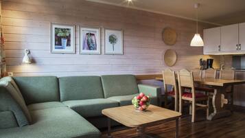 A modern living room with a green sofa, a wooden dining table with chairs, and a small wooden coffee table with a fruit bowl. There are framed pictures on the wall and a pendant light above the table.