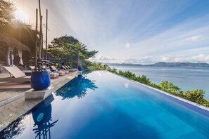 A infinity pool with a clear view of the sea and mountains, lounge chairs, and a thatched-roof structure.