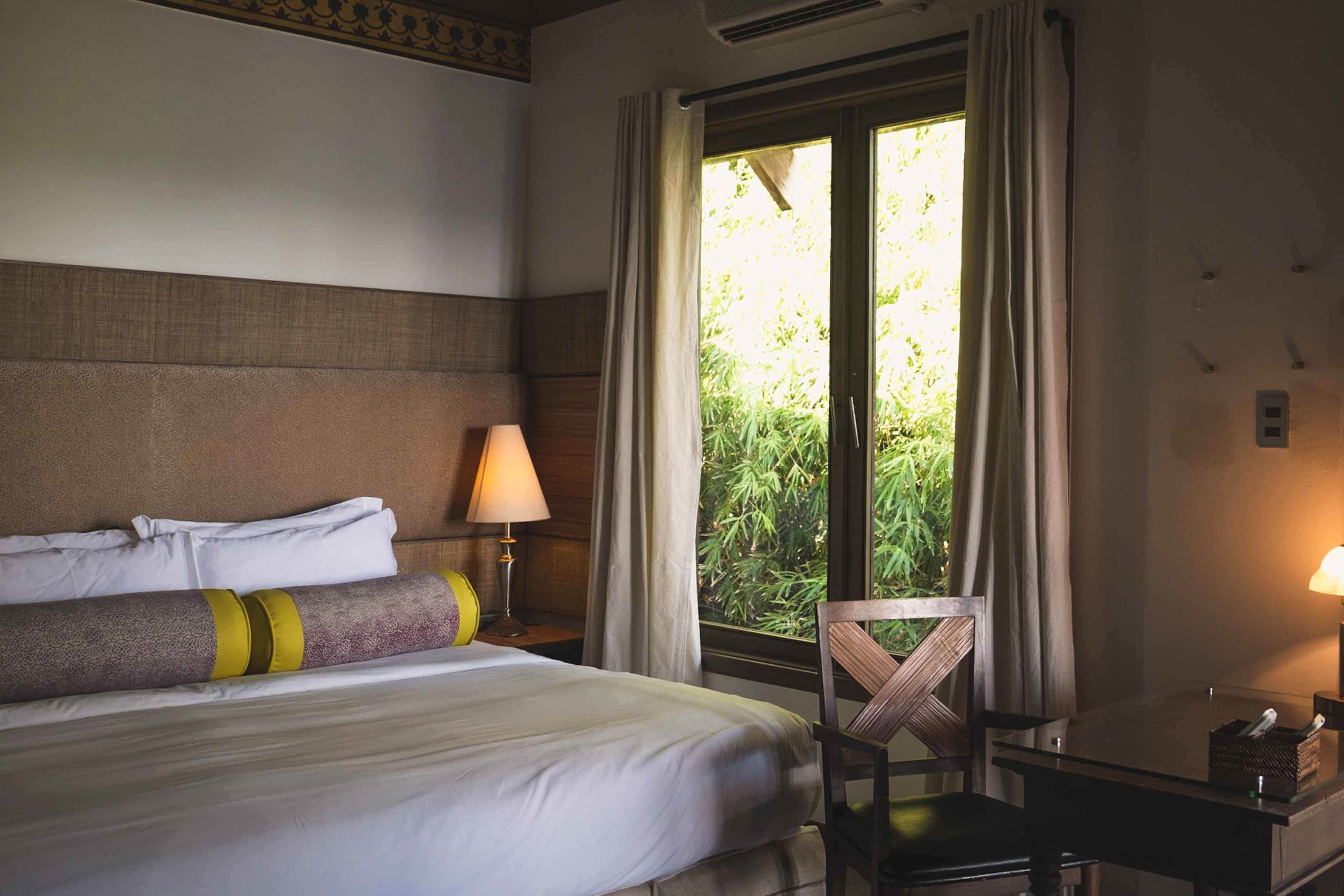 A hotel room with a bed, a desk, and a chair. There is a window with curtains and a view of greenery outside.