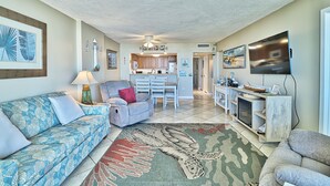 A living room with a sofa, a coffee table, a TV, and a kitchen in the background. -  Label Day Weekend Now Available 1st Floor-XXL Oceanfront Balcony OC Tower South (Myrtle Beach)
