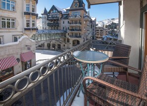 A balcony with wicker chairs and a glass table, overlooking a snow-covered street with historic buildings and a mountain in the background.