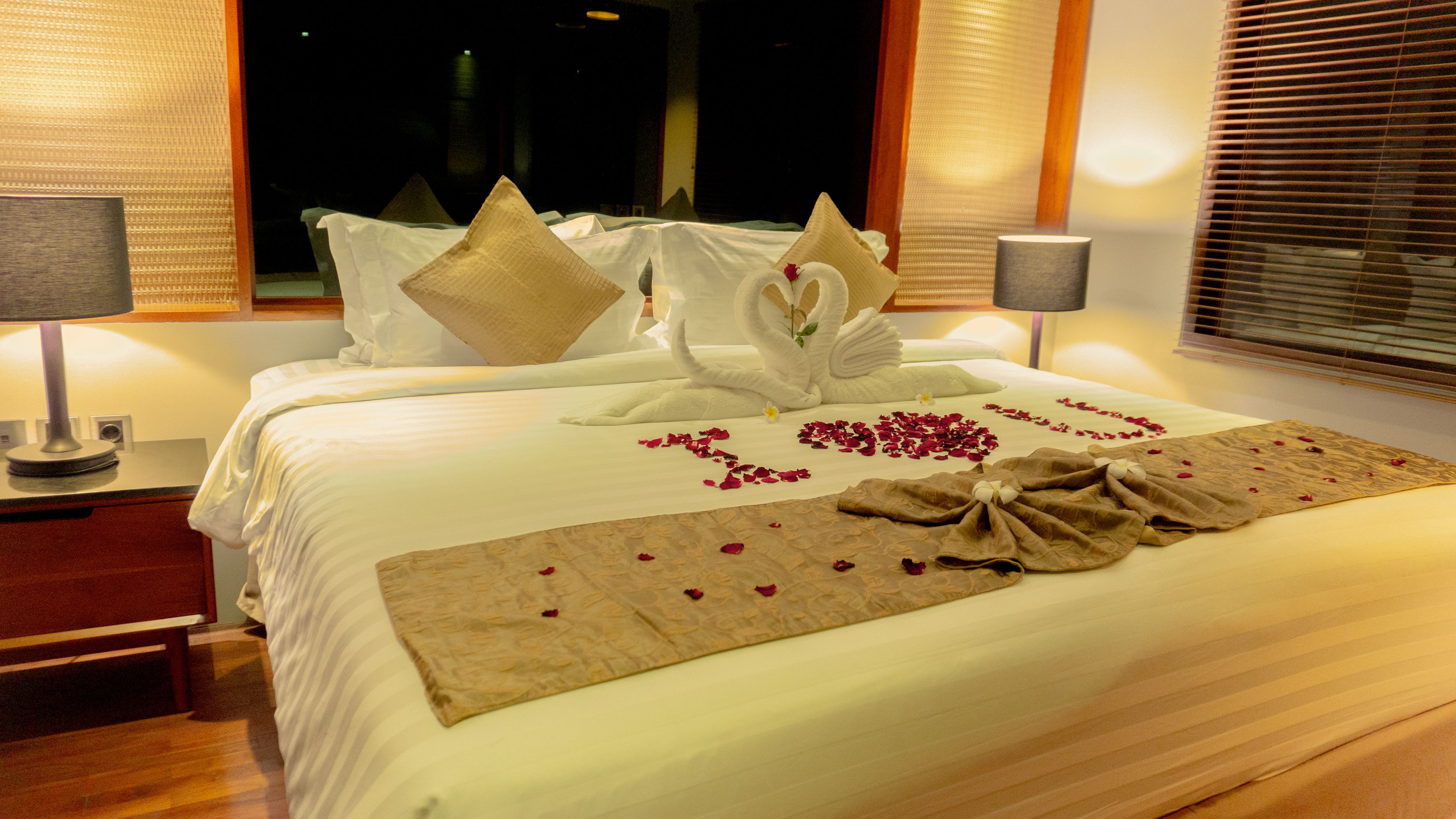 Una cama bien hecha con sábanas blancas, dos almohadas, un arreglo decorativo de toallas y pétalos de rosa sobre el edredón.
