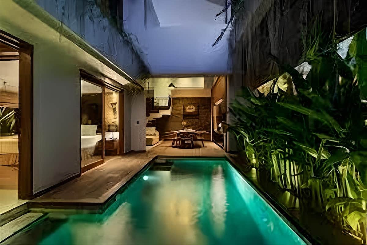 A modern indoor pool area with a wooden deck, a seating area with a table and chairs, and lush green plants.