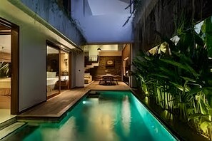 A modern indoor pool area with a wooden deck, a seating area with a table and chairs, and lush green plants. - Agranusa Signature Villa (Nusa Dua)