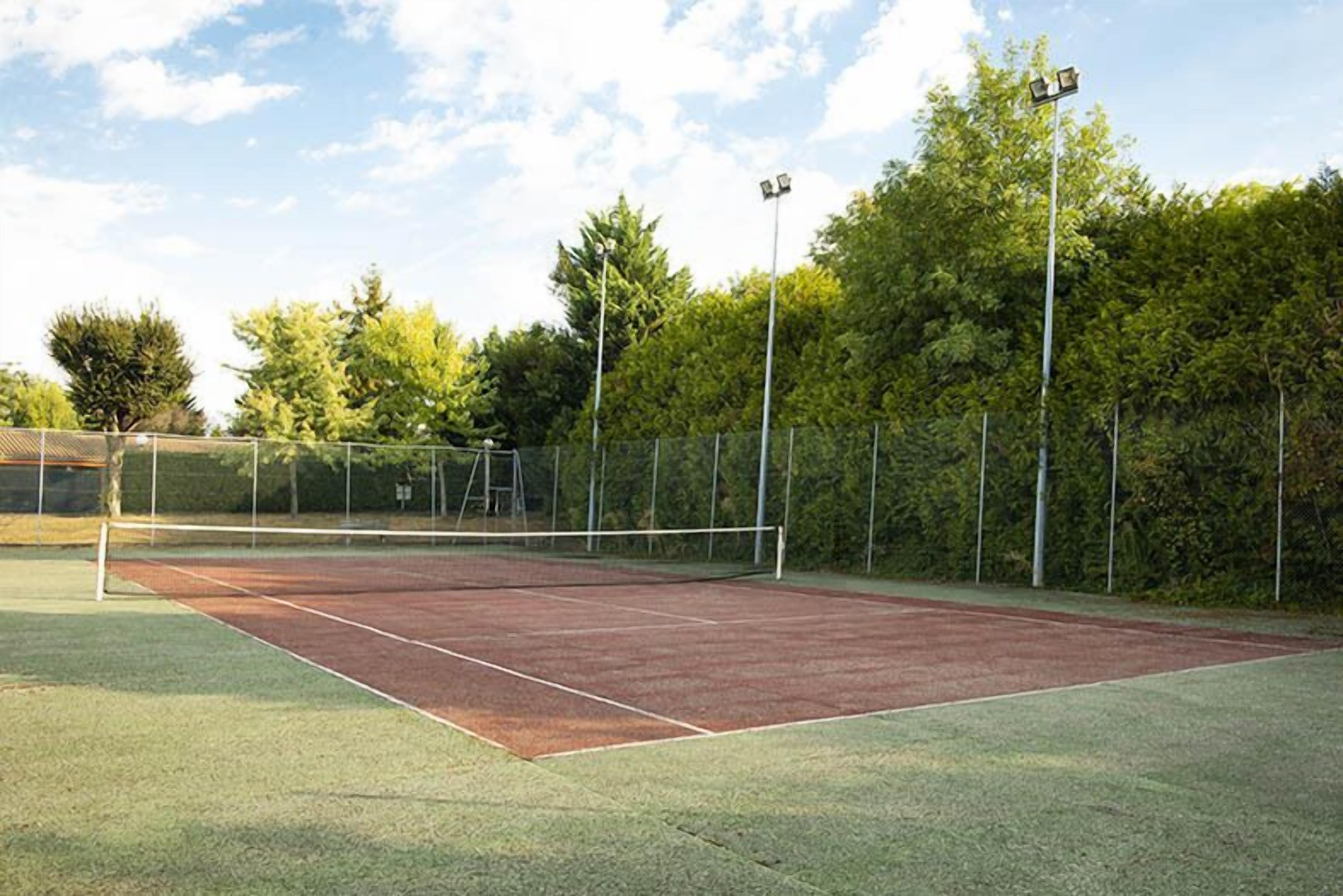 An outdoor tennis court with a net, surrounded by a fence and trees.