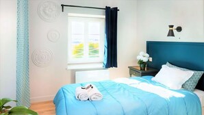 Een opgemaakt bed met een blauw beddegoed en witte kussens, een nachtkastje met een bloemenvaas, en een raam met gordijnen.