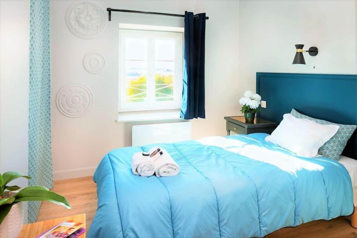 Ein ordentlich bezogenes Bett mit blauer Bettdecke und weißen Kissen, ein Nachttisch mit Blumen in einer Vase sowie ein Fenster mit Vorhängen.