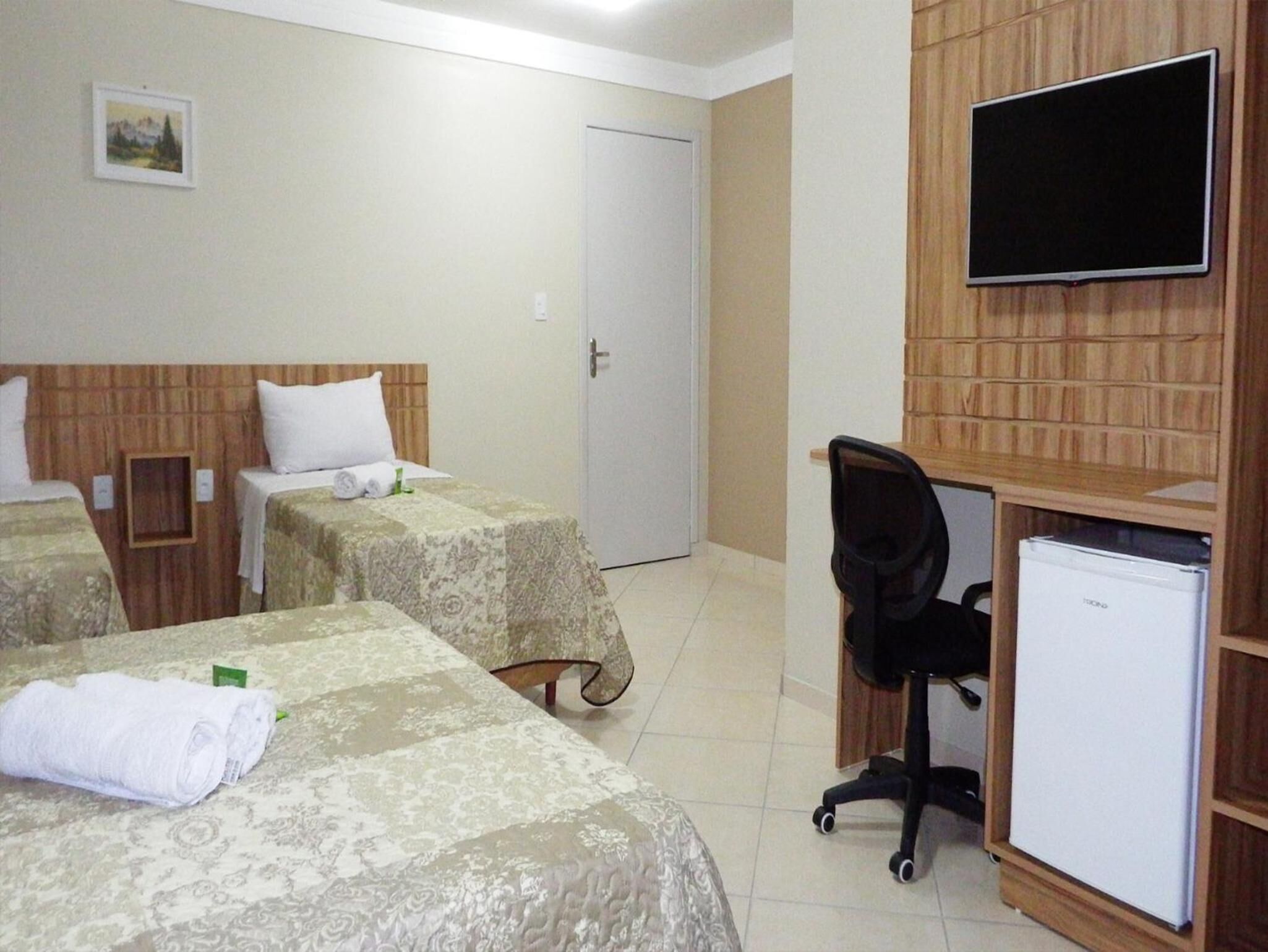 A hotel room with two beds, a desk, a chair, a TV, and a refrigerator.