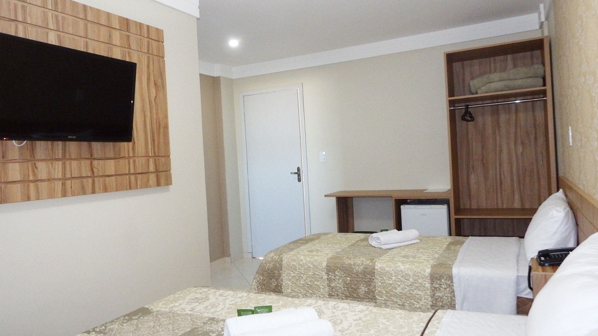 Een hotelkamer met twee bedden, een aan de muur bevestigde televisie, een kledingkast en een bureau met een stoel.
