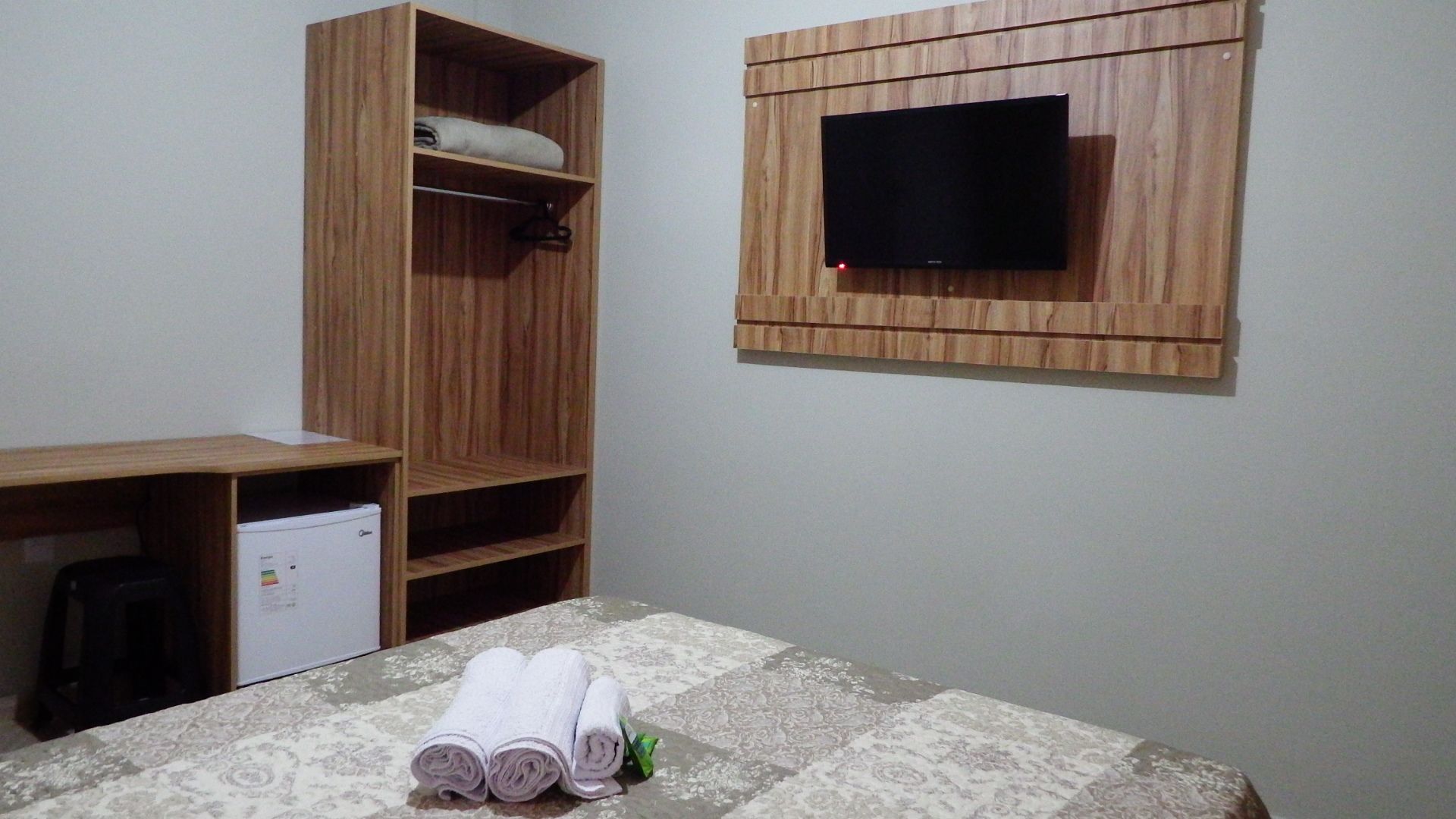 Quarto com cama, guarda-roupa de madeira, televisão na parede e um pequeno eletrodoméstico branco.