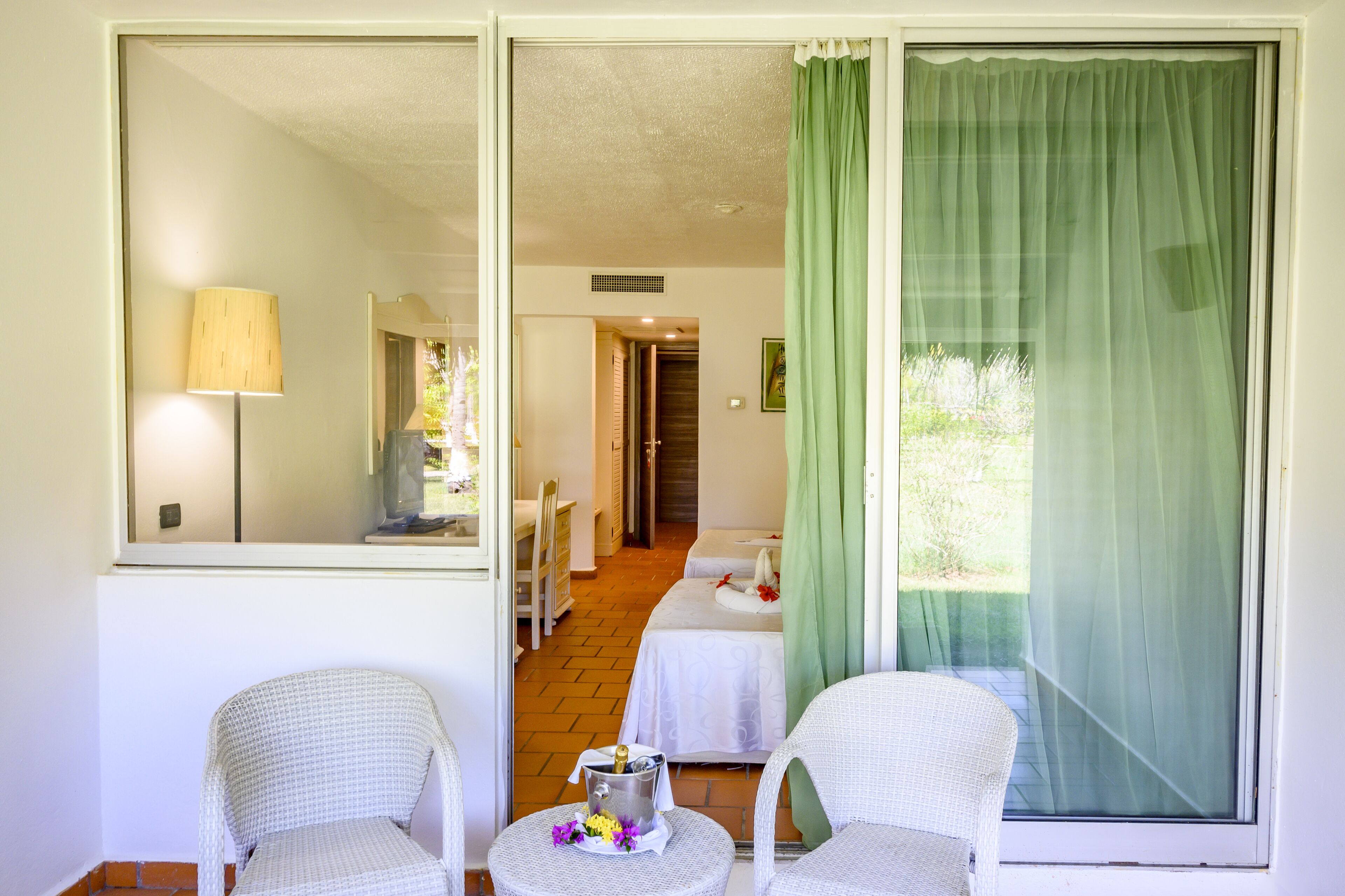 A hotel room with a balcony, a bed, a small table with a bottle and flowers, and a chair.