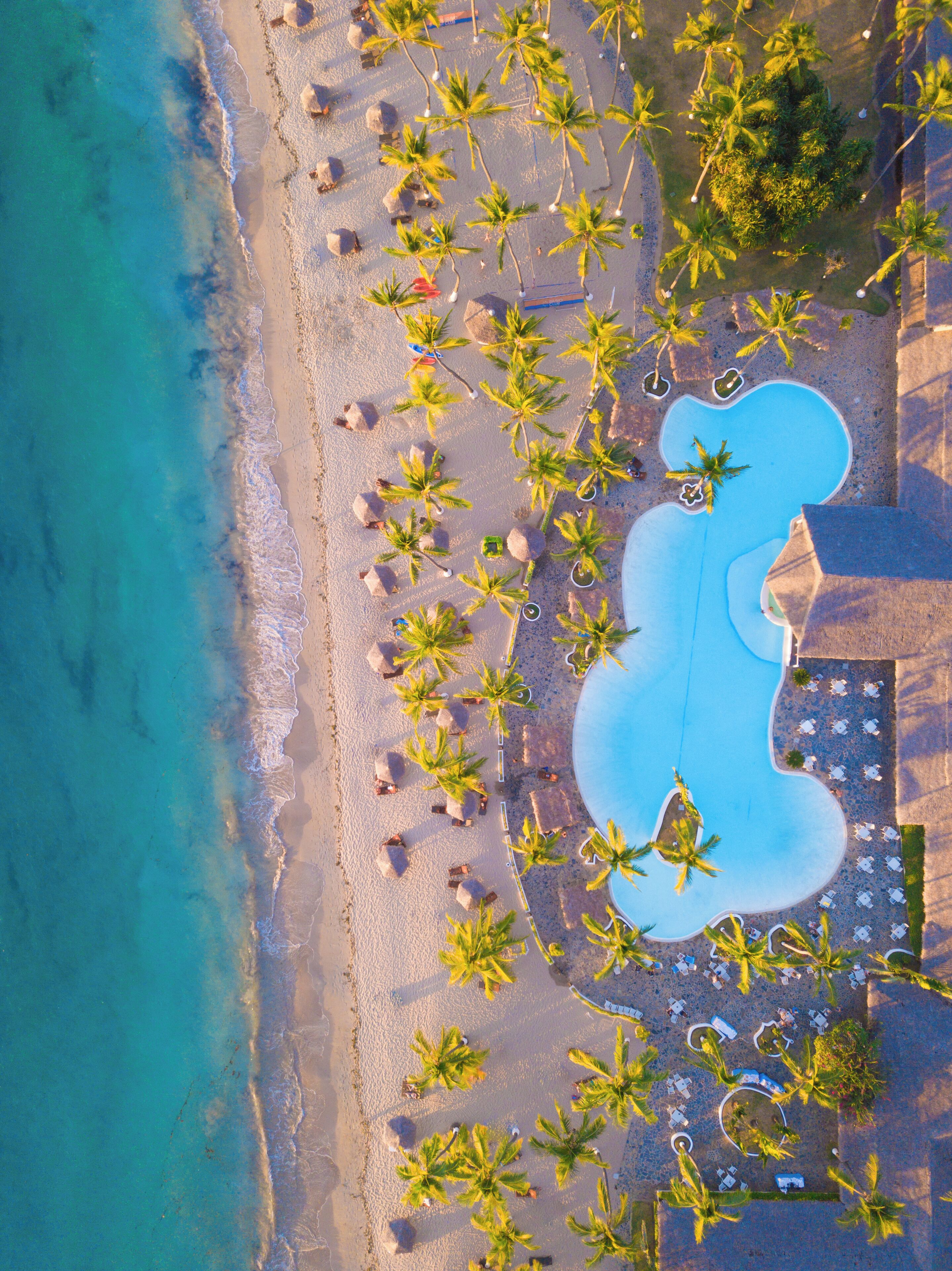 Vista aérea de un complejo turístico con piscina, palmeras y sillas de playa.