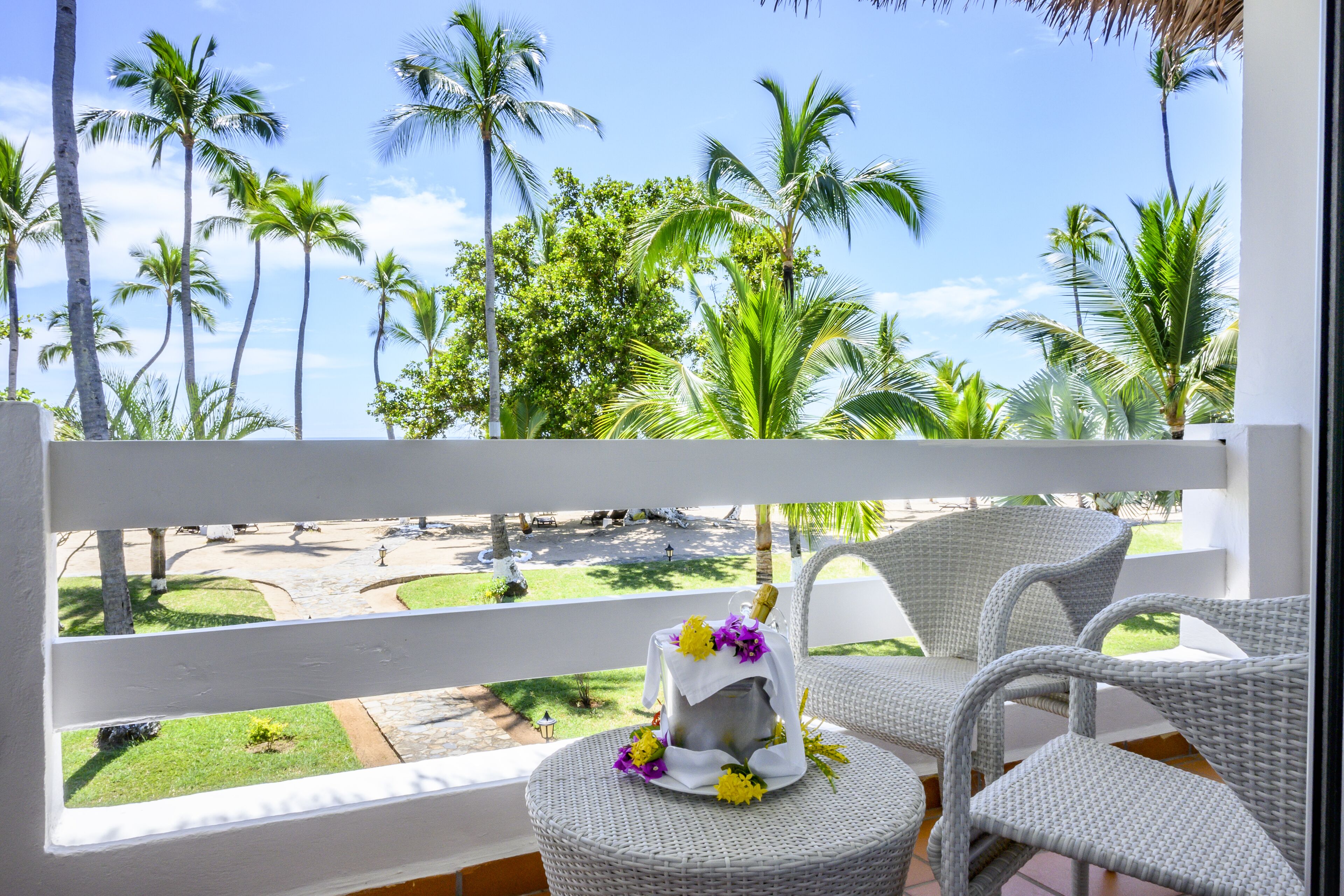 Un balcon avec des fauteuils en osier, une petite table agrémentée d’un objet décoratif, et une vue sur des palmiers et une piscine.