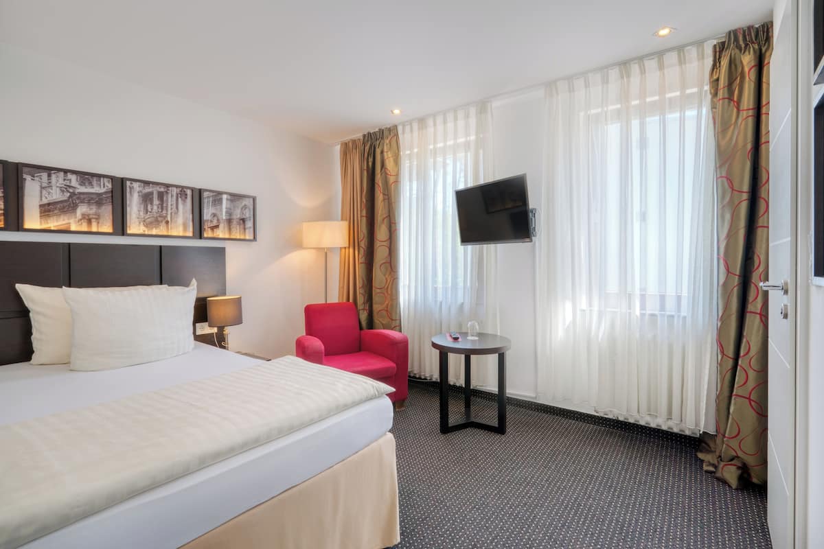 A hotel room with a bed, a red armchair, a small table with a lamp, and a TV mounted on the wall.