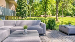 Ein moderner Außenbereich mit einer grauen Couch und passendem Hocker, einem kleinen Tisch mit einer Pflanze sowie einem gepflegten Garten mit Bäumen und Hecken.