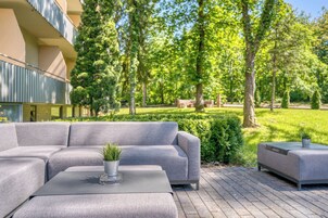 A modern outdoor seating area with a grey sofa and matching ottoman, a small table with a plant, and a well-maintained garden with trees and hedges.