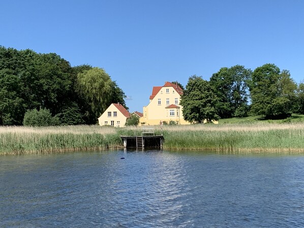 A house with a red roof and a dock extending into a calm body of water, surrounded by lush greenery. - Gut Grubnow Rügen (Neuenkirchen)