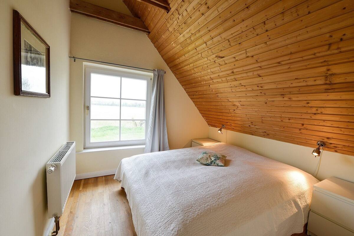 Une chambre avec un plafond en pente, une grande fenêtre avec des rideaux, un radiateur, un lit avec un oreiller et une table de chevet avec une lampe.