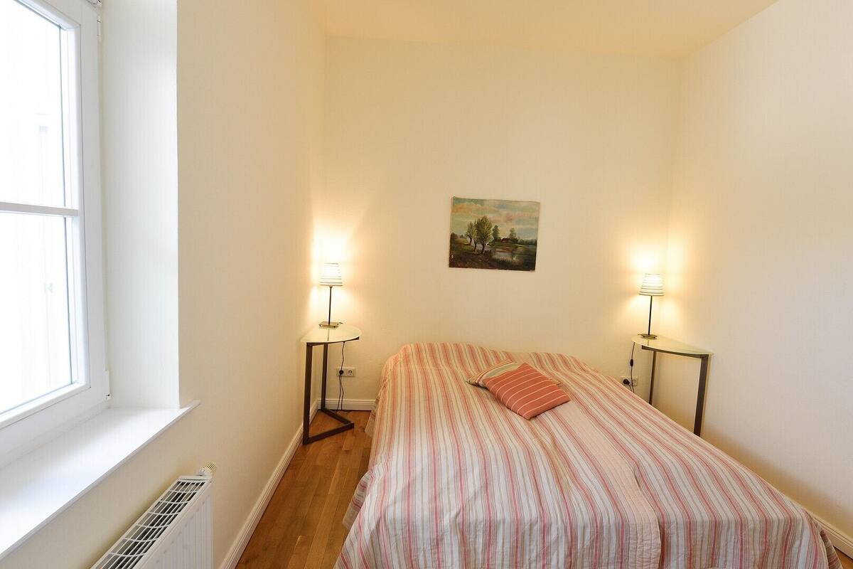 Une petite chambre bien éclairée, avec un lit simple, deux lampes de chevet, un tableau au mur et une fenêtre laissant entrer la lumière naturelle.