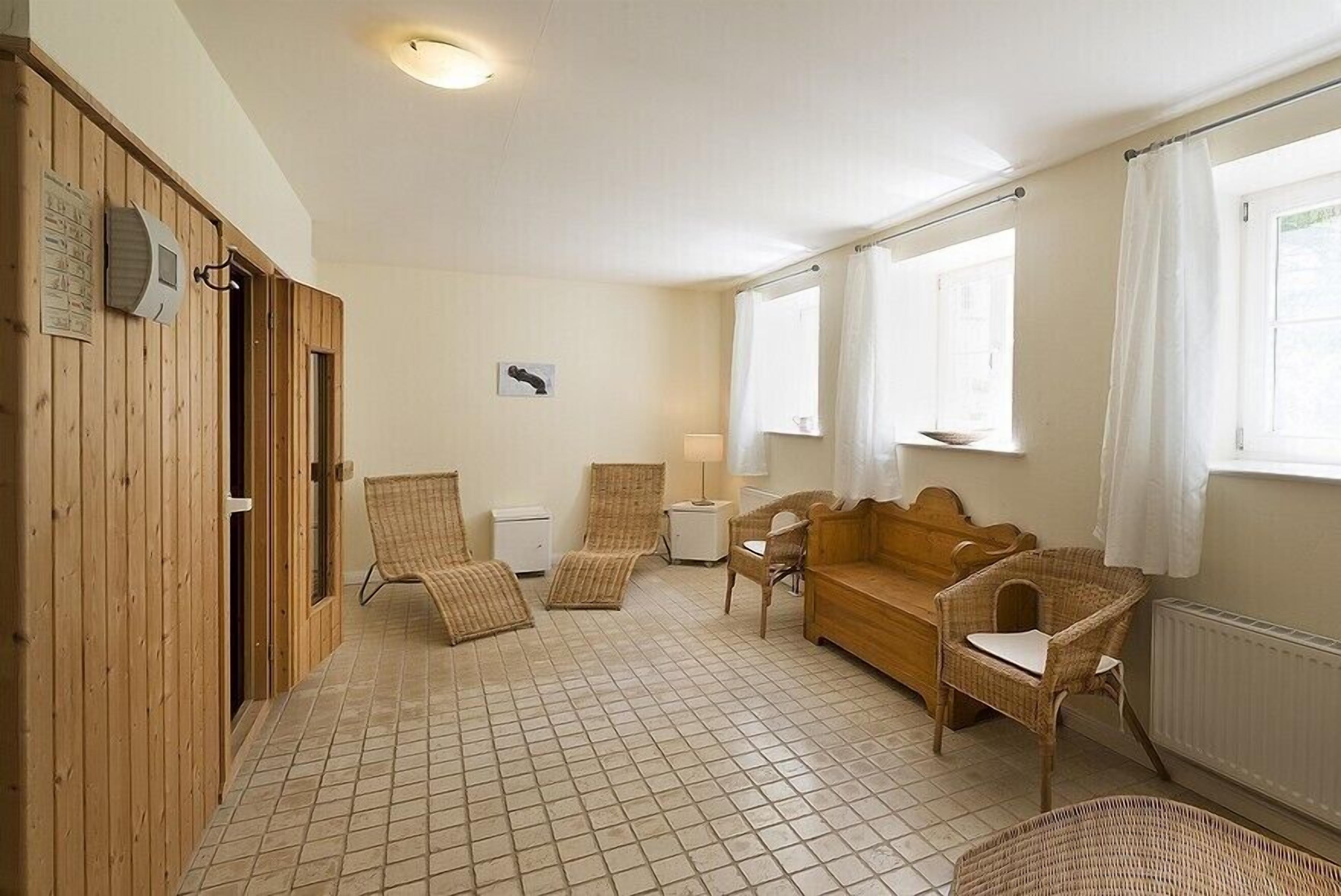 Una habitación con suelo de madera, un sofá de madera, dos sillas de mimbre, una mesita y una ventana con cortinas.