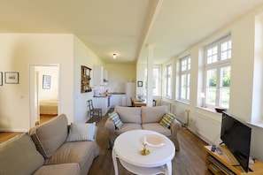 Un salón espacioso con un conjunto de sofás, una mesa de comedor y un televisor. Se observa una cocina al fondo, con armarios blancos y una ventana.