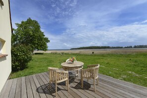 Una terraza de madera con una mesa redonda y cuatro sillas, con vistas a un área verde con un árbol y la playa.