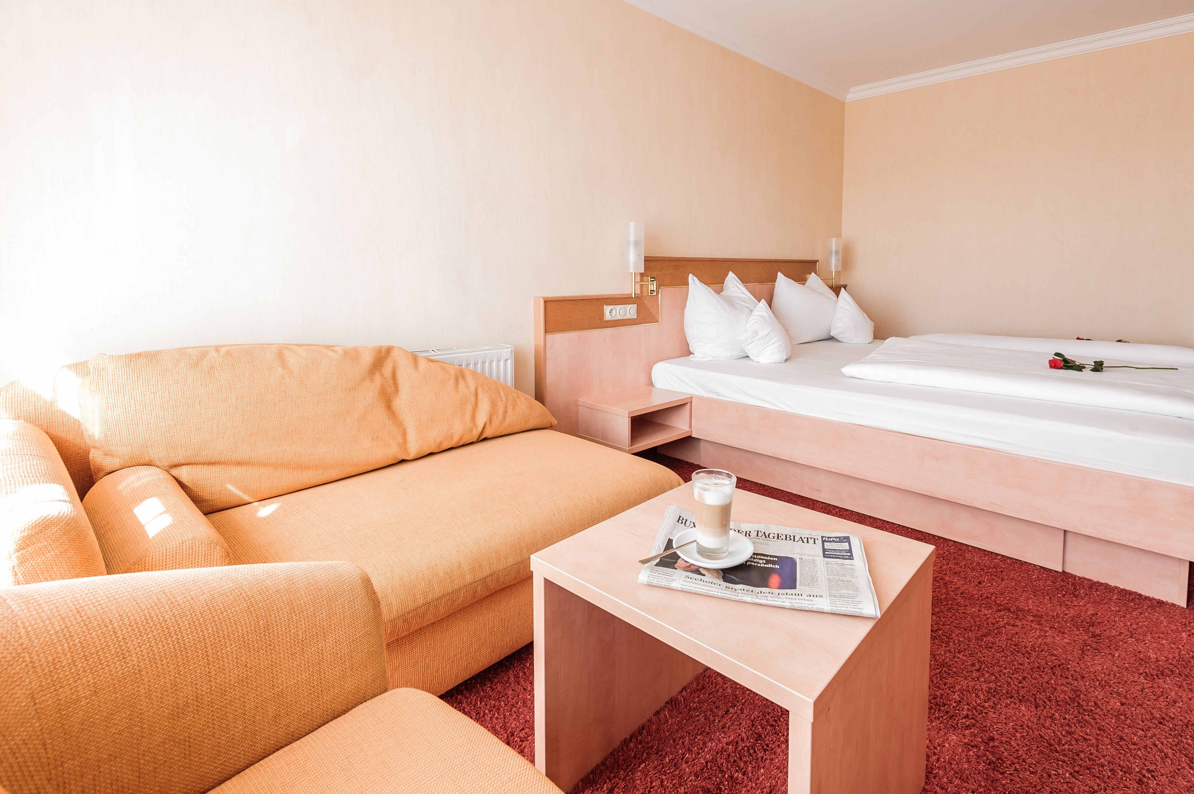A hotel room with a sofa, a bed, a small table with a newspaper and a cup, and a radiator.