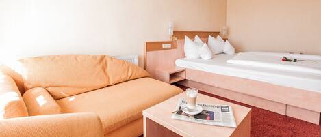 A hotel room with a sofa, a bed, a small table with a newspaper and a cup, and a radiator.