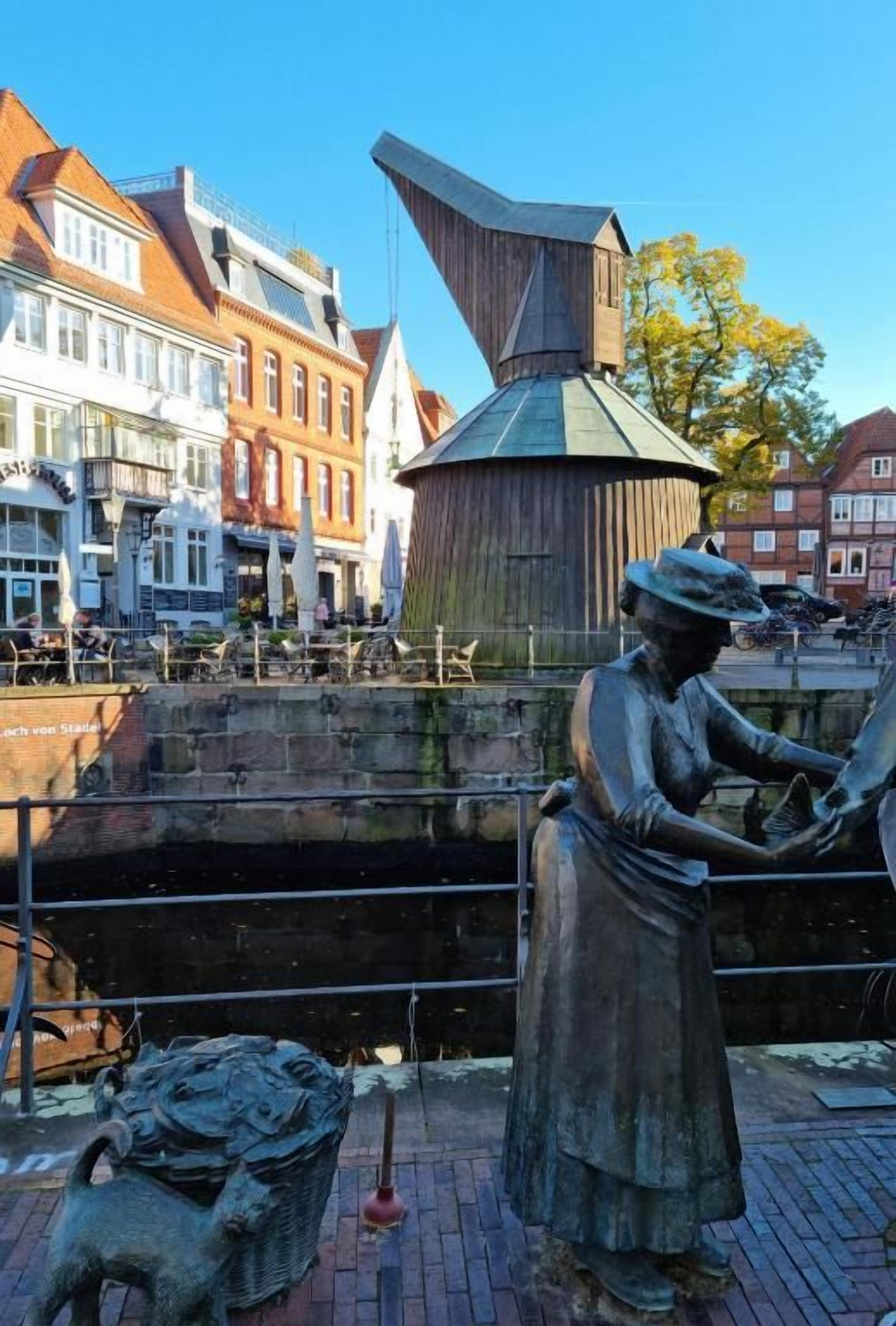 Une statue représentant une femme en jupe et portant un chapeau, une structure en bois et un canal avec des bâtiments en arrière-plan.