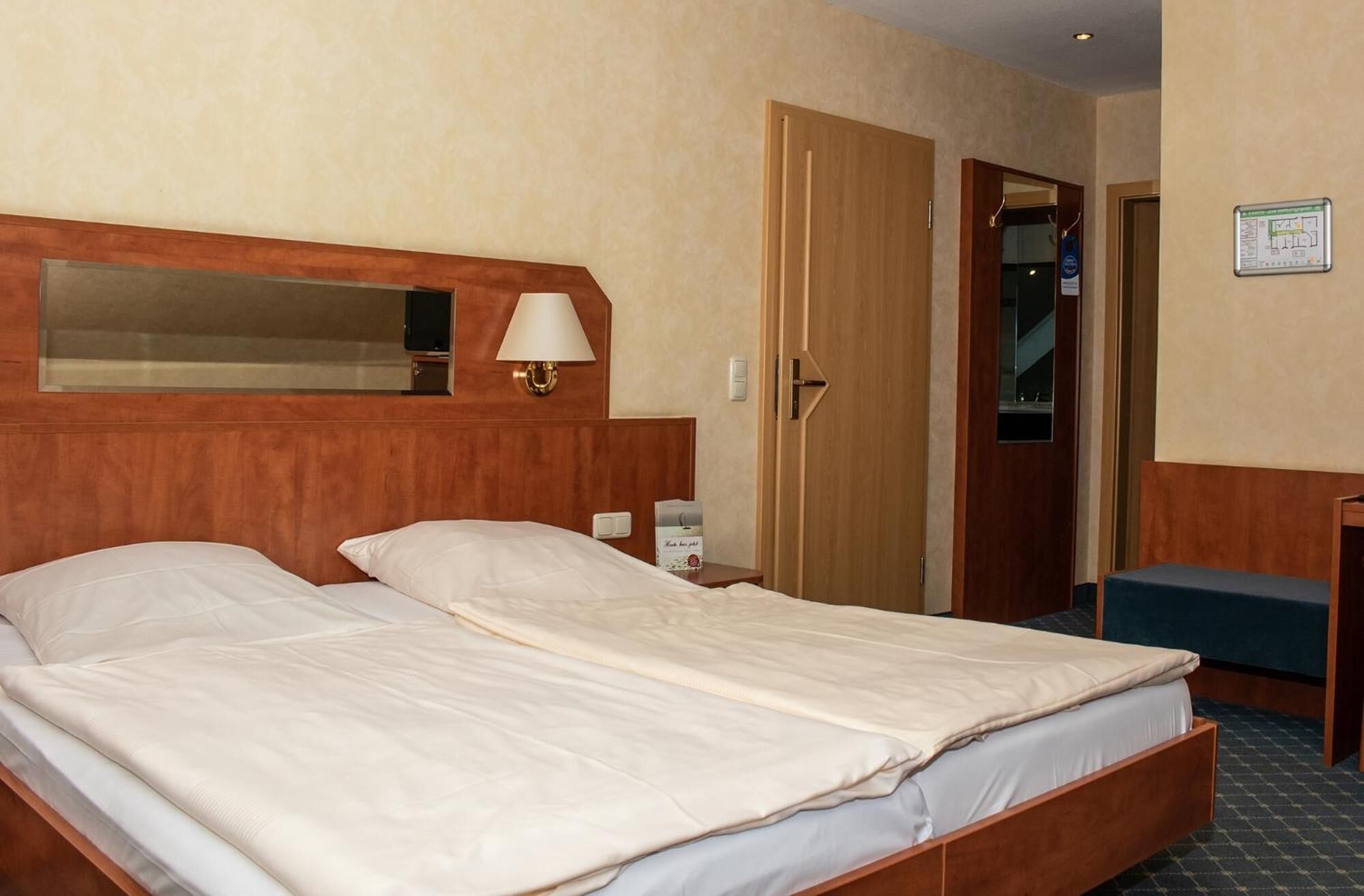 A hotel room with a double bed, a bedside table, a lamp, a mirror, a door, and a small desk with a chair.
