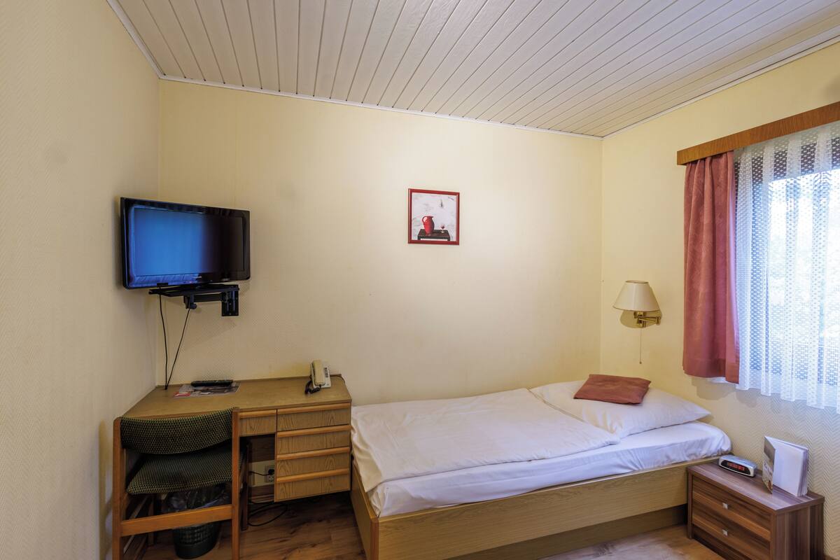 A small, neatly arranged bedroom with a single bed, a desk, a chair, a television, and a window with curtains.