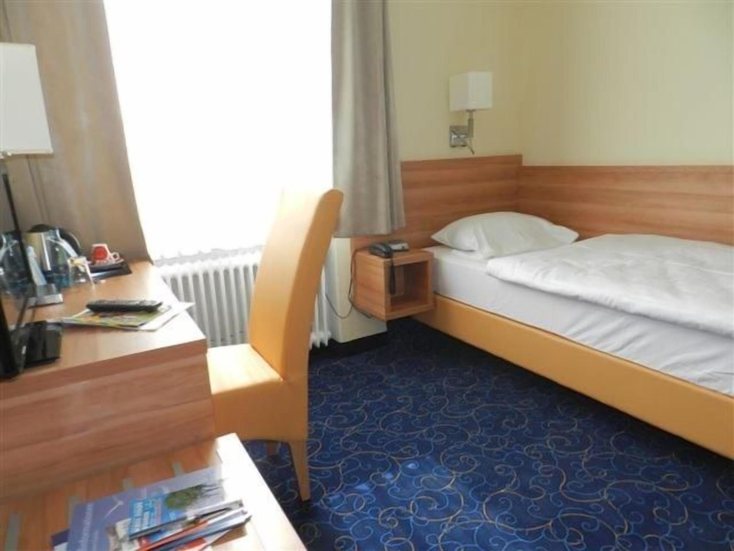 Ein Hotelzimmer mit einem Einzelbett, einem Schreibtisch, einem Stuhl und einem kleinen Tisch mit einer Lampe.