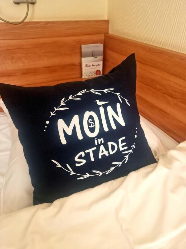 Una cama con una almohada que muestra el texto 'MOIN in STADE' y una tarjeta pequeña en la cabecera.